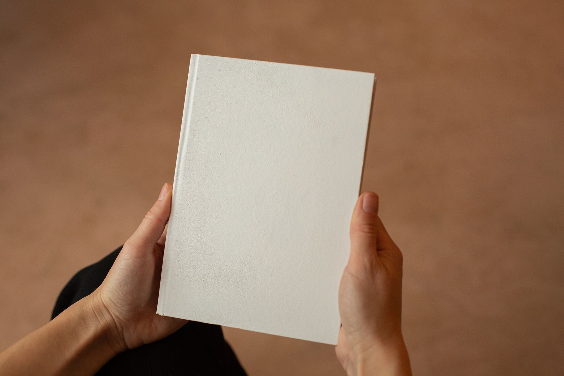 Two hands hold a blank warm-white hardback notebook